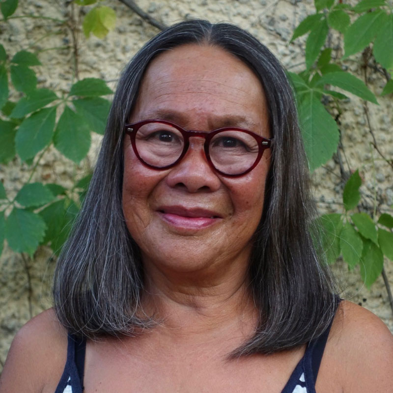 Jeannie Mah - Portrait of Indigenous woman smiling and wearing glasses. Behind her is a plant climbing on a wall.
