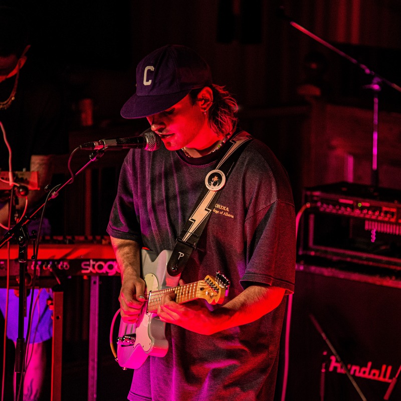Riley McLennan - Young man on stage in red lighting playing a guitar. He is wearing a cap, medium length hair.