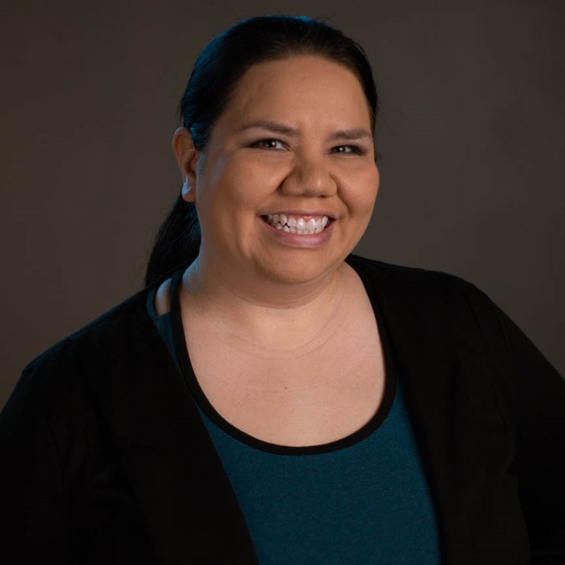 Jennifer Dawn Bishop - Portrait of Indigenous woman smiling. She is wearing s black cardigan and blue blouse.