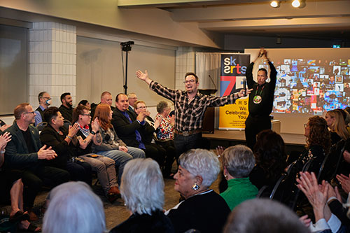 SK Arts 75th anniversary (2023) celebration at MacKenzie Art Gallery. Photo us of host Brad Bellegarde centre-isle of a seated, cheering crowd performing, In the background is the SK Arts banner and a pixelated screen of artists.