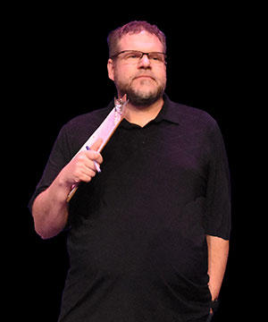 Ray Deans by Lyle Johnson for SK Arts Arts of Celebration 2025. Man in a black t-shirt and glasses holding a clipboard and a pen on stage.
